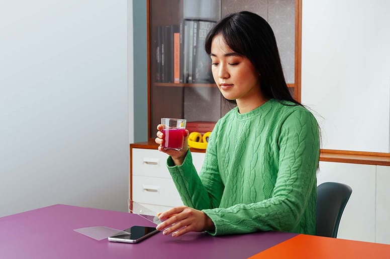 Junge Frau mit einem Glas Saft in der Hand sitzt vord dem Smartphone und vergleicht Panzerglas mit Displayschutzfolie