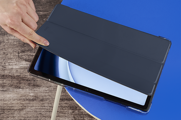 Auf einer blauen, runden Tischplatte liegt das Hama Tablet-Case "Fold Clear". Eine Hand kommt aus der linken oberen Ecke und klappt den Deckel des Covers auf, wodurch das aktivierte Display des Samsung Galaxy Tab 9 sichtbar wird.