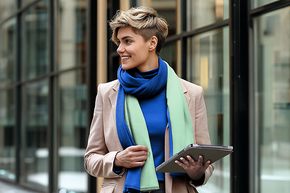 Eine junge Frau ist in einer Einkaufsstraße unterwegs und trägt ihr Tablet in der Hama Tablet-Hülle "Fold Clear" mit sich. Sie ist frühlingshaft gekleidet mit einem beigen Blazer, blauem T-Shirt und grün-blauem Schal.