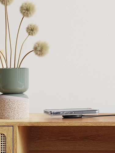 Das kabellose Qi2-Ladegerät mit induktivem Ladepad von Hama liegt auf einem hölzernen Sideboard, ein Smartphone liegt darauf und wird geladen. Danaben stehen zwei Vasen in Pastelltönen mit Trockenblumen darin.