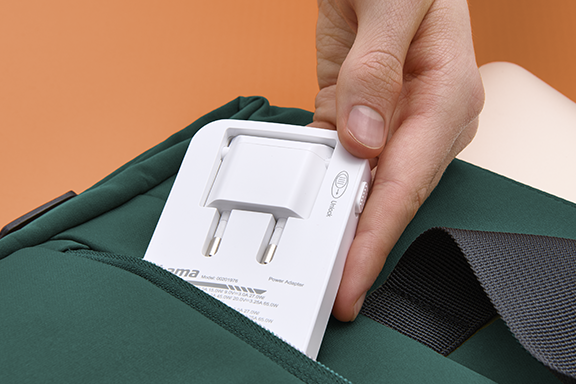 Beim Hama Schnellladegerät kann der Netzwerkstecker eingeklappt werden, sodass es noch platzsparender verstaut werden kann. Im Bild wird es von einer Hand in einen grünen Rucksack gesteckt.