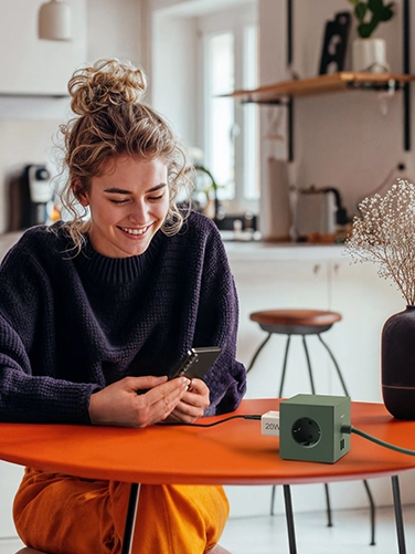 Junge Frau sitzt an einem Esstisch und bedient ihr Smartphone, während dieses mit dem Hama Schnellladegerät, 20W geladen wird