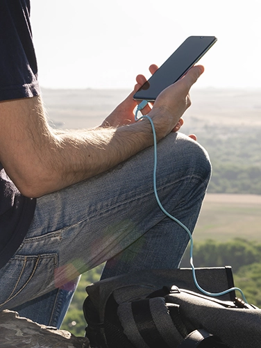 Mann sitzt beim Wandern in auf einem Hügel und lädt sein Smartphone mit dem Hama Power Pack "PD10-HD"