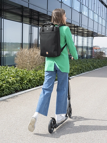 Eine junge fährt auf einem E-Scooter vom Betrachter weg und hat den Hama Laptop-Rucksack "Extreme Protect" auf dem Rücken.