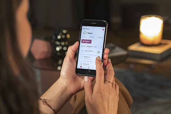 Eine junge Frau sitzt in einem dunklen Wohnzimmer und steuert die Temperatur im Raum über die Hama Smart Home-App.