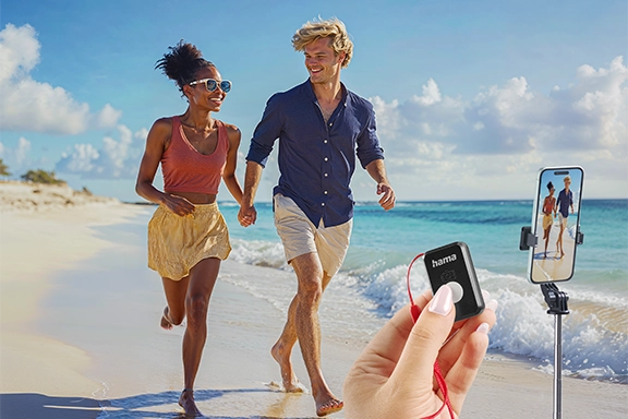 Im Hintergrund läuft ein junges Pärchen gut gelaunt und händchenhaltend am Sand-Strand entlang. Im Vordergrund sind freigestellte Bilder vom Stativ "Fancy Stand 70 II" und einer Hand, die die Bluetooth-Fernbedienung hält, zu sehen